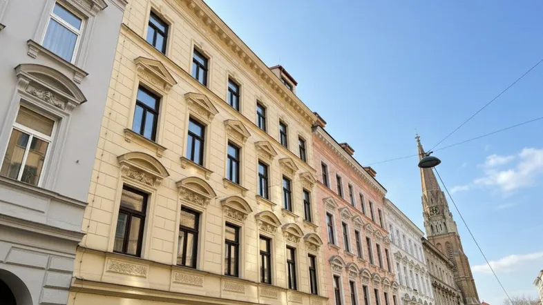 Historische Fassade eines Wohngebäudes in einer belebten Straße mit Kirchturm im Hintergrund.