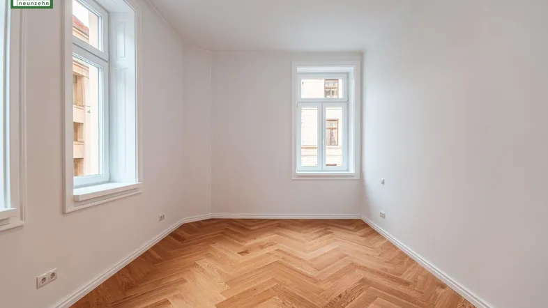 Geräumiges, helles Zimmer mit elegantem Fischgrätparkett und mehreren Fenstern, bereit zur individuellen Gestaltung.