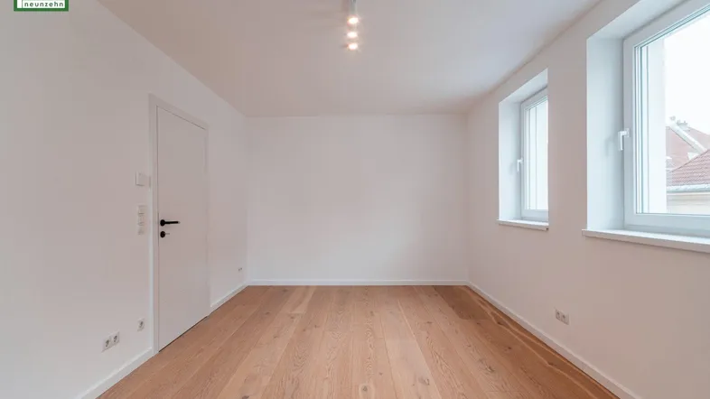 Helles, leeres Zimmer mit Holzboden und zwei Fenstern, bereit zur individuellen Gestaltung.