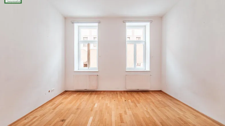 Helles, leeres Zimmer mit zwei Fenstern, Parkettboden und Heizkörpern, bereit zur individuellen Gestaltung.