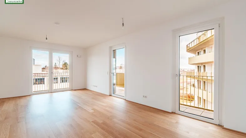 Helles, leeres Zimmer mit Holzboden und Zugang zu mehreren Balkonen in einem Neubau.