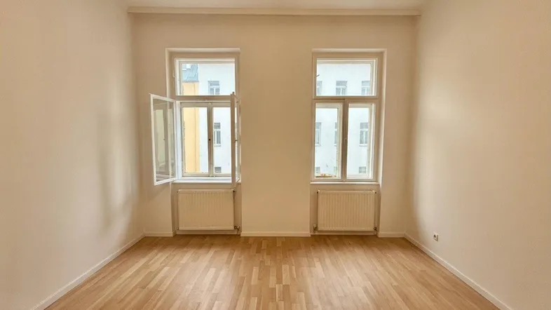 Helles Zimmer mit zwei großen Fenstern, Parkettboden und Heizkörpern, ideal als Wohn- oder Schlafzimmer.