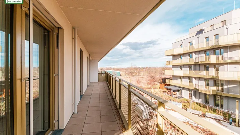 Langer Balkon mit Fliesenboden und Blick auf moderne Wohngebäude in sonniger Umgebung.