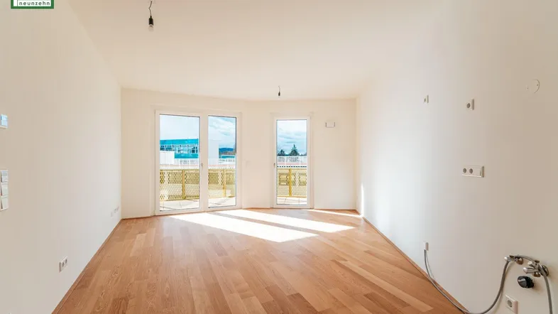Helles, leeres Wohnzimmer mit Holzboden und Zugang zu einem sonnigen Balkon.