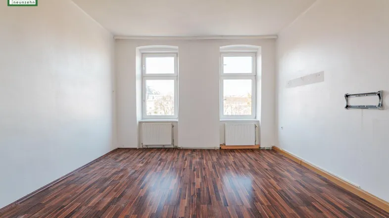 Helles, leeres Zimmer mit dunklem Laminatboden und zwei Fenstern, bereit zur individuellen Gestaltung.