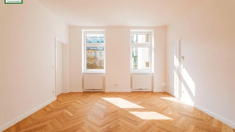 Helles, leeres Zimmer mit Fischgrätparkettboden und zwei Fenstern, das viel Tageslicht hereinlässt.