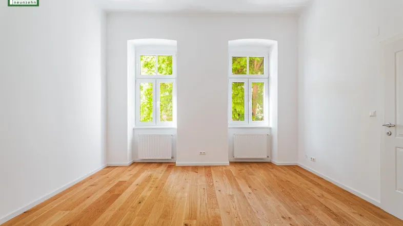 Helles, leeres Zimmer mit frisch verlegtem Holzparkett und zwei Fenstern mit Blick ins Grüne.