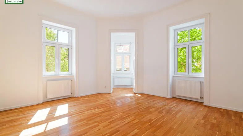 Helles, leeres Zimmer mit Parkettboden und großen Fenstern, die viel Tageslicht hereinlassen.