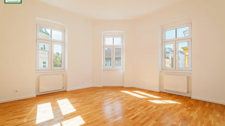 Helles, leeres Zimmer mit Fischgrätparkett und großen Fenstern, durch die Sonnenlicht fällt.
