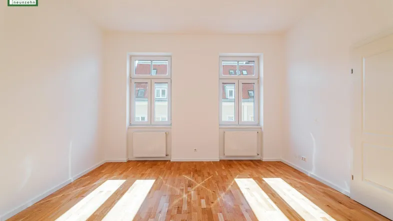 Helles Zimmer mit Holzboden und zwei Fenstern, das viel Tageslicht hereinlässt.