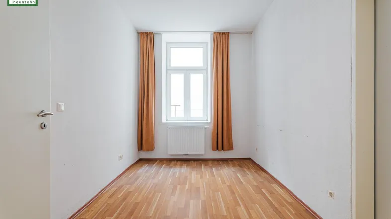 Helles Zimmer mit Holzfußboden, weiß gestrichenen Wänden und einem Fenster mit Heizkörper.