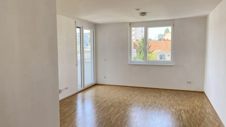Geräumiges, leeres Wohnzimmer mit Holzboden und Zugang zu einem Balkon.