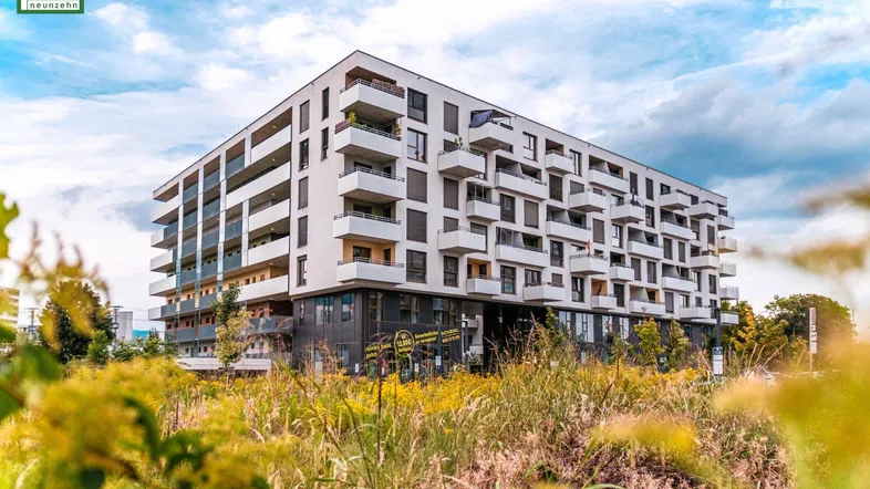 Modernes Mehrfamilienhaus mit vielen Balkonen und grüner Umgebung unter bewölktem Himmel.