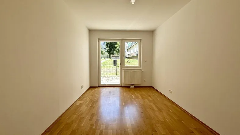 Helles, leeres Zimmer mit Parkettboden und Zugang zu einem grünen Außenbereich.