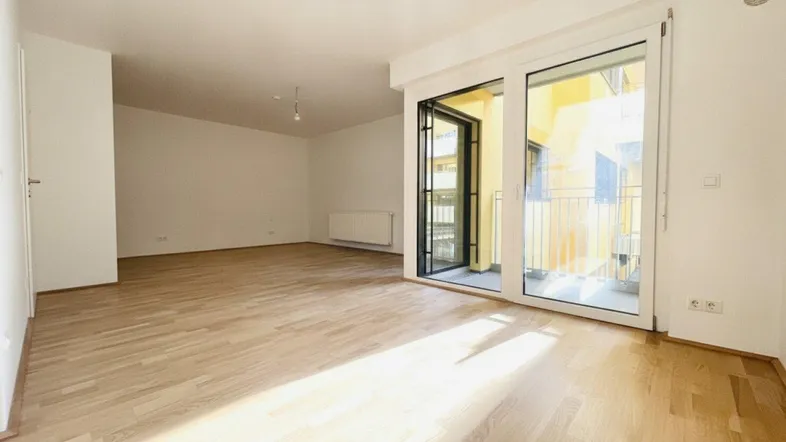 Helles, leeres Zimmer mit Parkettboden und Zugang zu einem Balkon.