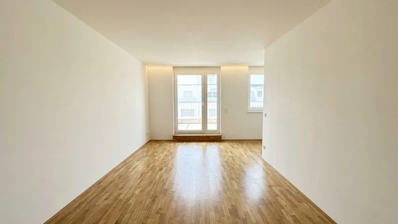 Helles, leeres Wohnzimmer mit Holzboden und Zugang zu einem Balkon durch große Türen.
