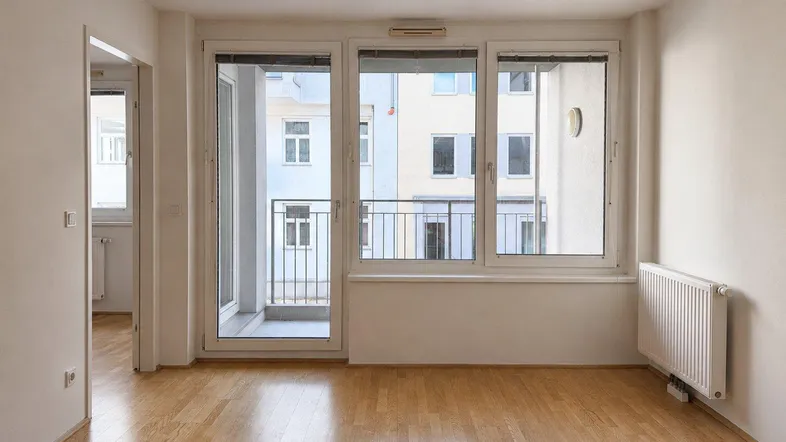 Geräumiger Wohnbereich mit Parkettboden, großen Fenstern und Zugang zu einem französischen Balkon.