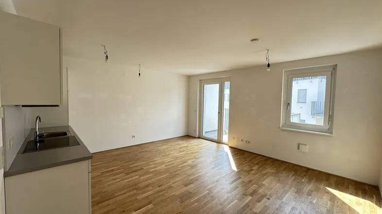 Helles Zimmer mit Holzboden, Balkonzugang und einer kleinen Küchenzeile.