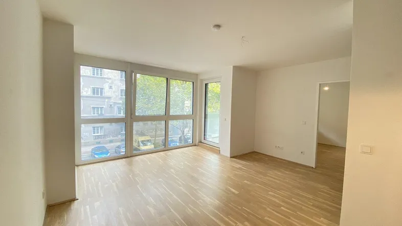 Helles Wohnzimmer mit großen Fenstern, Parkettboden und Zugang zu weiteren Räumen.