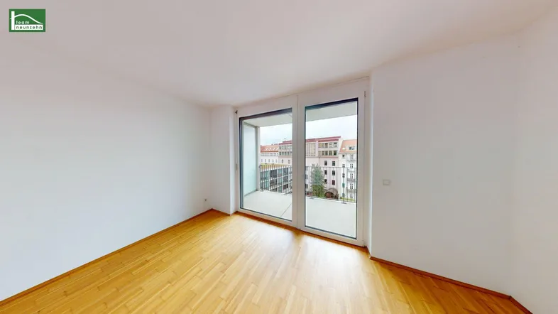 Helles Zimmer mit Holzboden und großen Fenstern, die auf einen Balkon führen.