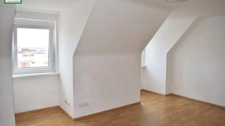 Helles Zimmer im Dachgeschoss mit Dachschrägen, Parkettboden und Blick auf die Umgebung.