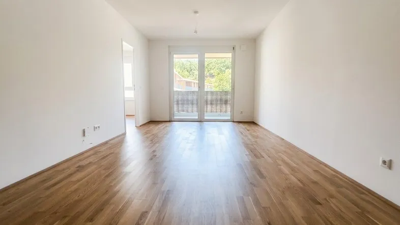 Helles, leeres Zimmer mit Holzboden und Zugang zu einem Balkon mit Blick ins Grüne.