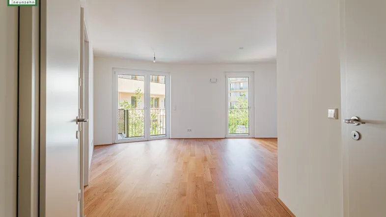 Helles, leeres Wohnzimmer mit Holzboden und Zugang zu zwei französischen Balkonen.