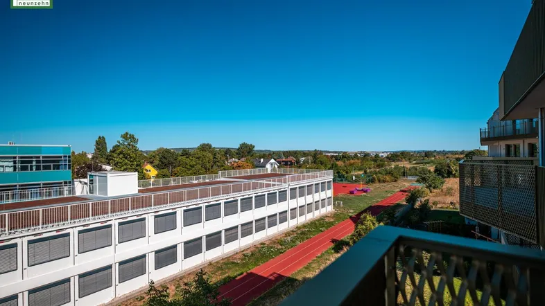 Weitblick vom Balkon über eine Sportanlage und grüne Landschaft unter blauem Himmel.