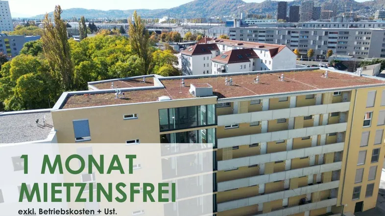 Luftaufnahme eines modernen Mehrfamilienhauses mit Balkonen und begrüntem Flachdach in städtischer Umgebung mit Bergblick.
