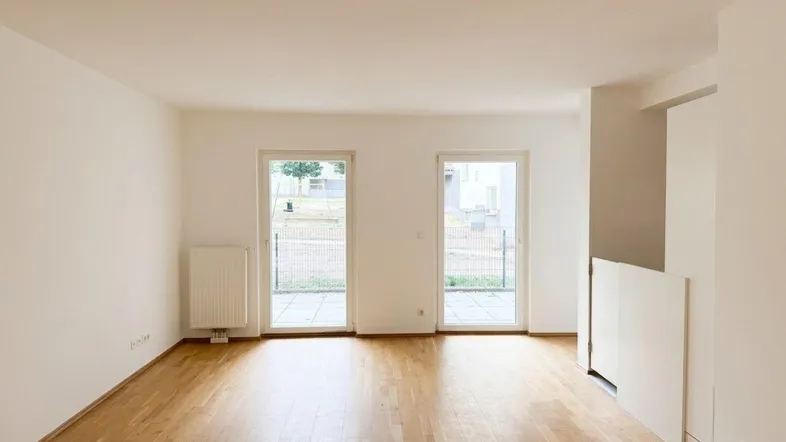 Helles, leeres Wohnzimmer mit Parkettboden und Zugang zu einer Terrasse oder einem Balkon.