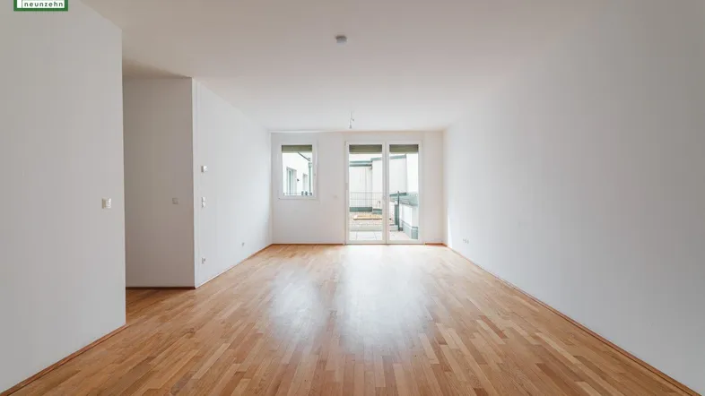 Helles, leeres Wohnzimmer mit Parkettboden und Zugang zu einem Balkon oder einer Terrasse.