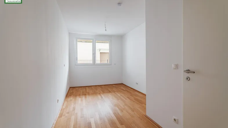 Helles, leeres Zimmer mit Holzparkettboden und einem Fenster, das viel Tageslicht hereinlässt.