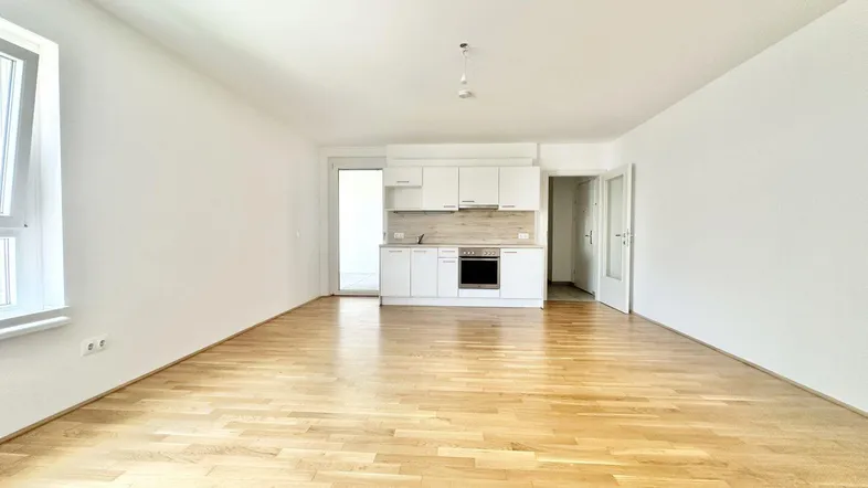 Heller, leerer Wohnbereich mit offener Küchenzeile, Parkettboden und Zugang zum Balkon.