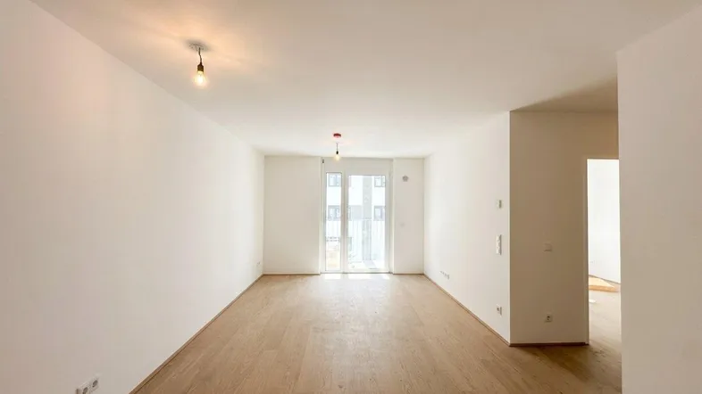 Helles, leeres Wohnzimmer mit Holzboden und Zugang zu einem Balkon oder einer Terrasse.