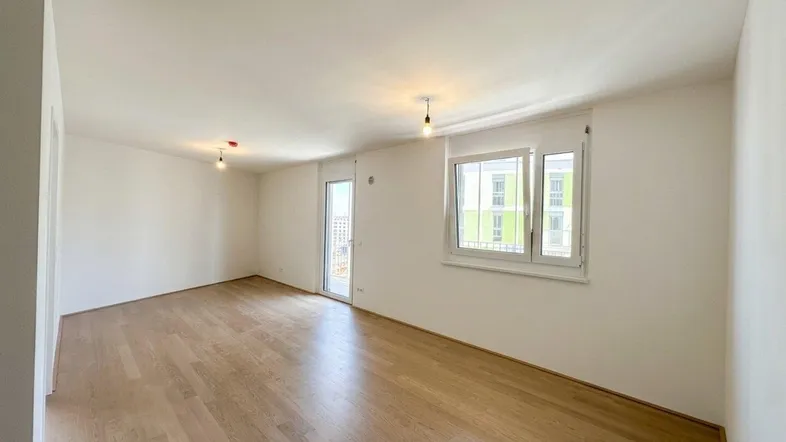 Helles, leeres Wohnzimmer mit hellem Parkettboden und Zugang zu einem Balkon.