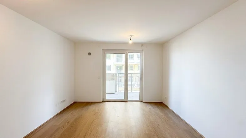 Helles, leeres Zimmer mit Holzboden und Zugang zu einem Balkon in einem Neubau.
