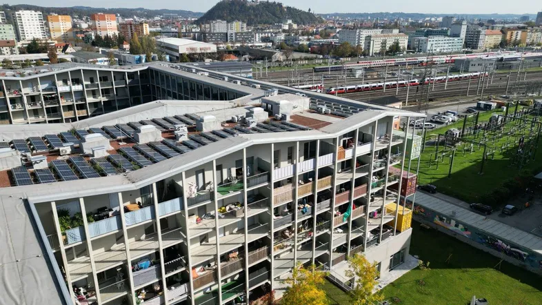 Luftaufnahme eines modernen Wohnkomplexes mit vielen Balkonen und Solaranlagen auf dem Dach, umgeben von Stadtlandschaft.