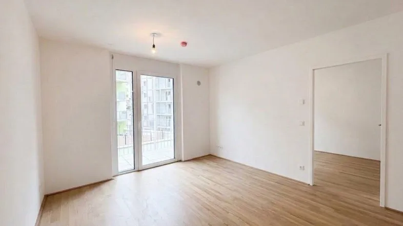 Helles, leeres Zimmer mit Holzboden und Zugang zu einem Balkon in einem Neubau.