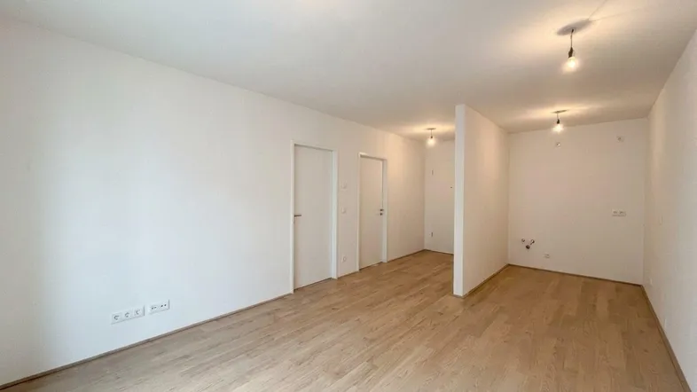 Geräumiges, leeres Zimmer mit hellem Holzboden und mehreren Türen in einem modernen Gebäude.