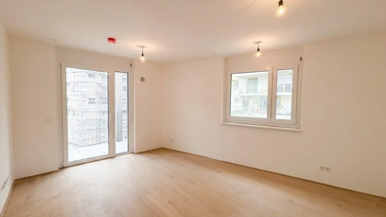 Helles, leeres Zimmer mit Holzboden, großen Fenstern und Zugang zu einem Außenbereich.