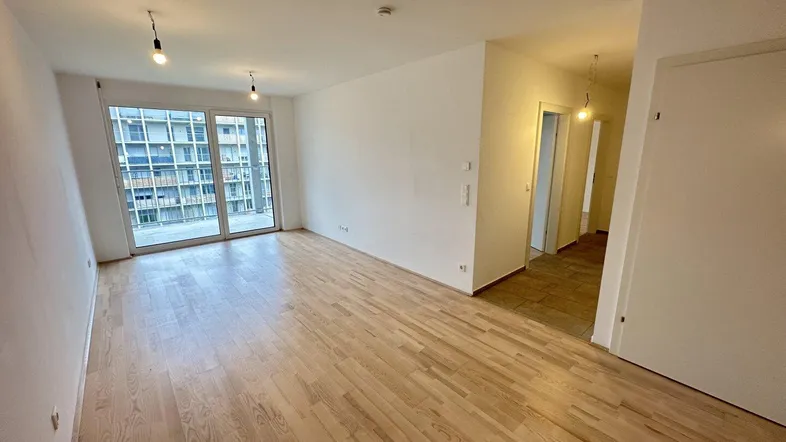 Helles, leeres Wohnzimmer mit Holzboden und Zugang zu einem großen Balkon.