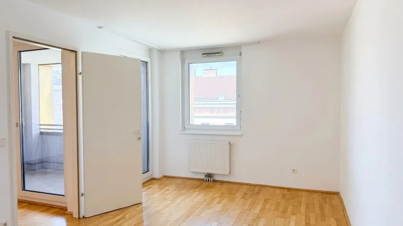 Helles Zimmer mit Parkettboden, weißen Wänden, Fenster und Zugang zu einem Balkon.