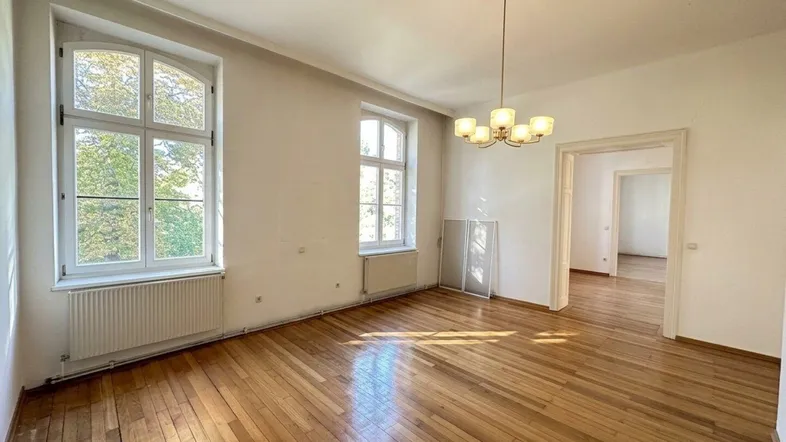 Helles, geräumiges Wohnzimmer mit hohen Fenstern, Parkettboden und Zugang zu weiteren Räumen.