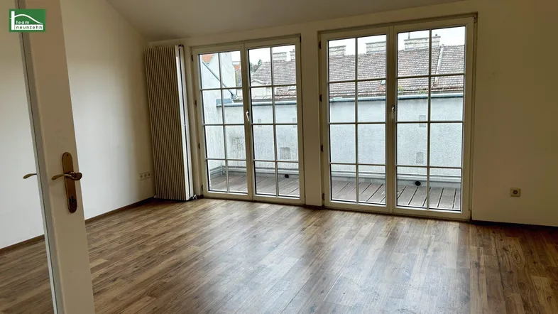 Helles Zimmer mit Holzboden und großen Fenstern, die auf einen Balkon führen.
