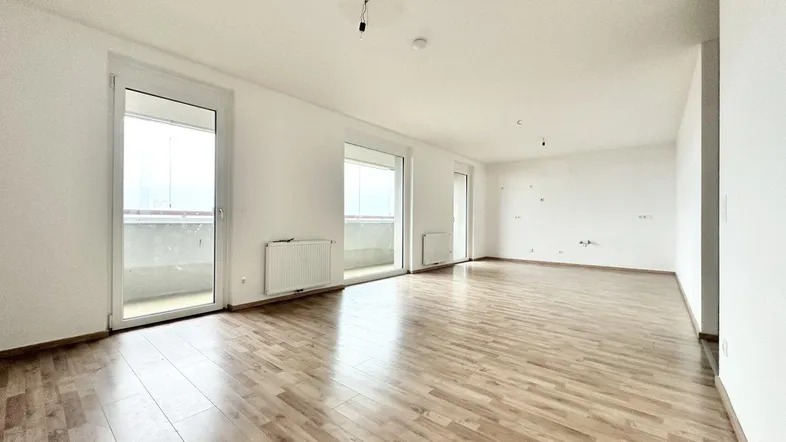 Helles, leeres Wohnzimmer mit Laminatboden und Zugang zu einem Balkon.