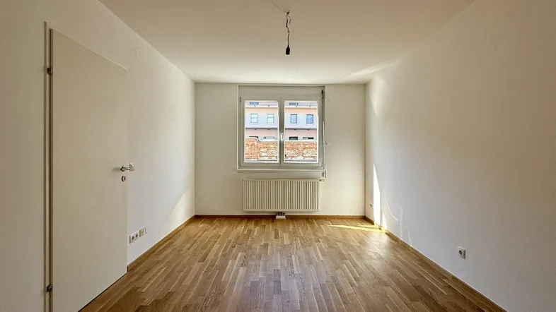 Helles, leeres Zimmer mit Holzparkettboden und einem Fenster mit Heizkörper darunter, bereit zur Nutzung.