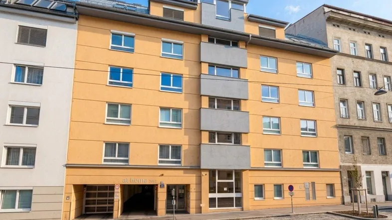 Mehrstöckiges Wohngebäude mit gelber Fassade und grauen Balkonen an einer Stadtstraße.