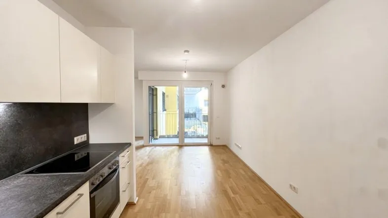 Heller offener Wohnbereich mit integrierter Küchenzeile, Parkettboden und Zugang zum Balkon.
