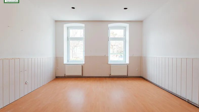Heller, leerer Raum mit zwei Fenstern, Holzvertäfelung an den Wänden und Laminatboden.