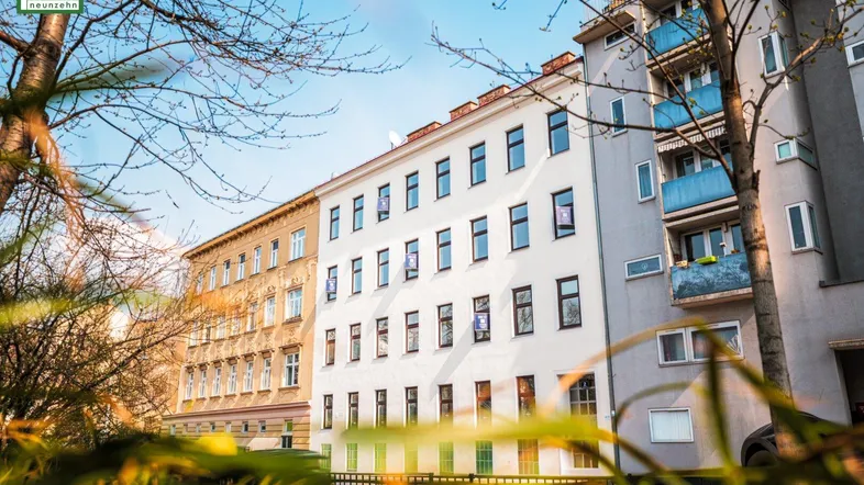 Helle Fassade eines Mehrfamilienhauses mit vielen Fenstern, umgeben von Bäumen bei Sonnenschein.